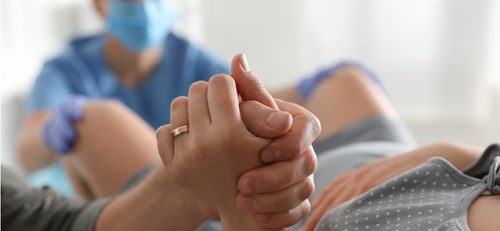 Hands held in the foreground, a woman lying down giving birth, in the background a medic with gown and mask