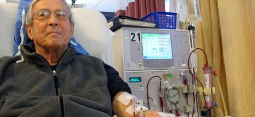 A man relaxed in a clinic chair beside a haemodialysis machine. The man has two tubes connected to his arm, carrying blood to and from the machine. The tubes are taped to his arm.