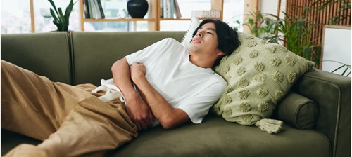 Young man lying on sofa clutching his abdomen, face showing discomfort