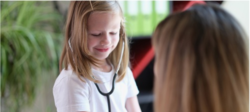 A girl of around 8-10 years smiles gently and looks down. She has on a stethoscope, in front of her we see the back of the head of a woman who appears to be communicating with the girl
