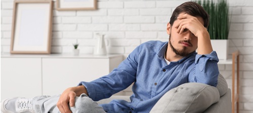 A young man with a beard, sitting on a grey sofa, head in one hand as if tired