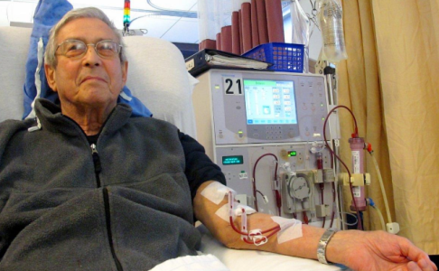 A man relaxed in a clinic chair beside a haemodialysis machine. The man has two tubes connected to his arm, carrying blood to and from the machine. The tubes are taped to his arm.