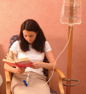 A photo of a woman sat in a chair reading a book while doing a dialysis fluid swap. A bag of fluid hangs from a stand. A tube from the bag connects to her peritoneal catheter, unseen under her t-shirt.
